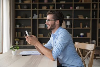 Gülümseyen 35 'lik işadamı, erkek ofis çalışanı modern akıllı telefon kullanarak çalışma masasında oturuyor, e-postaları kontrol ediyor, sms' de iyi haberler okuyor, iş ve iş için kolay teknoloji kullanımının keyfini çıkarıyor.