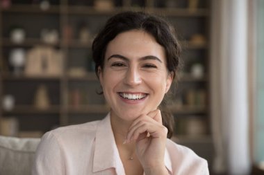 Geniş dişli gülümsemesi olan genç bir kadın kanepeye oturmuş kollarıyla çenesine dokunarak modern evde poz veriyor. Aşırı mutlu görünüyor. Mutlu 20 'ler kadın portresi konsepti