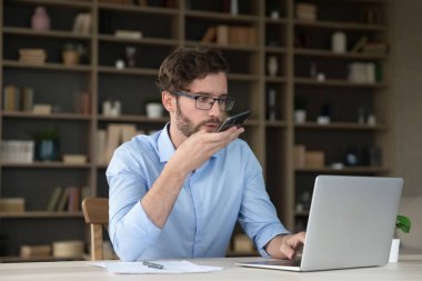 Ciddi genç adam masada oturur, akıllı telefon konuşmasını hoparlörden yapar, bilgiyi paylaşır, modern teknolojiyi kullanarak müşteriye profesyonel destek sağlar, sesli mesaj gönderir, çevrimiçi çeviri uygulaması kullanarak tercüme eder.