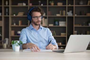 Kulaklıklı genç bir solak masa başına oturur dizüstü bilgisayara bakar, çevrimiçi öğrenir, ses kurslarını dinler, dil becerilerini geliştirir, bilgi alır, metin ödevlerini çevrimiçi okur.