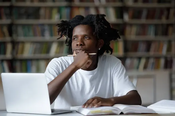 stock image Pensive African guy sit at table with laptop and books in library looking into distance, making task, thinking over solution, prepare for college or university exams. Studies, tech, education concept