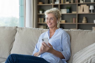 Çekici, yaşlı bir kadın, dijital yayın hizmetlerini kablosuz kulaklıkla kullanarak sesli mesajlar ya da favori müzik dinliyor. Kanepeye oturup, modern akıllı telefon aleti kullanıyor. Oturma odasında dinleniyor.