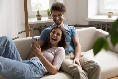 Neşeli genç çift internet üzerinden iletişim kurmak, dinlenmek, kanepede kucaklaşmak, birlikte eğlenmek, iyi haberler almak, gülümsemek, gülmek için akıllı telefondan başvuruyor.