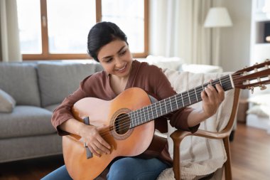 Gülümseyen Hintli kadın akustik gitar tutuyor, müzik besteliyor, evdeki rahat koltukta oturuyor, mutlu genç bayan müzisyen müzik aleti çalıyor, boş zamanların tadını çıkarıyor, hobi kavramı.