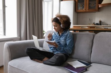 Modern kablosuz teknolojiyi kullanarak e-öğrenme, öğretmenle çevrimiçi sınıf, yeni bilgi edinme. Afrikalı genç kız kulaklık takıyor yabancı dil dersleri dinliyor, evdeki kanepeye oturtuyor.