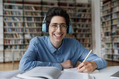 Kulaklık takan öğrenci kütüphanede oturmuş, yeni bilgiler alıyor, sınavlara teknoloji kullanarak hazırlanıyor, kameraya gülümsüyor. Video çağrısı etkinliği, eğitim, öğrenim kavramı