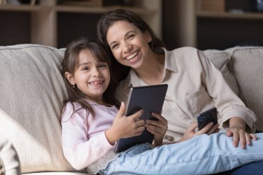 Mutlu kadın ve küçük kızı dijital tablet cihazıyla kanepede dinleniyor, gülümsüyor, kameraya bakıyor. Modern teknoloji, yeni uygulama, internet kullanıcıları portresi, boş zaman ve eğlence araçları kullanarak evde