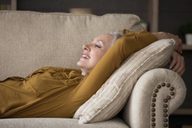 Mutlu, olgun bir kadın rahat bir kanepede uzanıyor, gülümsüyor, gülüyor, hayal kuruyor, başka yere bakıyor, temiz hava alıyor, vücudunu esnetiyor, dinleniyor, dinleniyor, rahatlıyor, rahatlıyor, rahatlıyor.