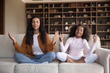 Afrikalı bir kadın. Anaokulu kızı oturma odasındaki rahat koltukta bacak bacak üstüne atmış halde oturuyor. Mudra jesti yaparak evde meditasyon yaptırıyor. Anne çocuğa iyi yaşam alışkanlığı, yaşam tarzı, yoga egzersiz konsepti öğretiyor.
