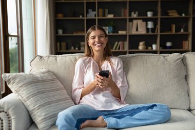 Genç nesil kadın portresi internette rahat bir koltukta oturup modern akıllı telefon tebessümüyle kameraya bakıyor. Ev hizmetlerinde teslimat, e-alışveriş mutlu müşteri, mobil uygulama kullanımı konsepti