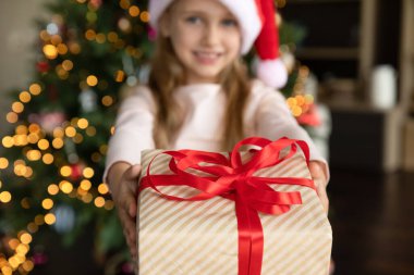 Küçük kızların Noel hediyesi kutusunu kapat. Tatlı anaokulu çocuğu kucaklıyor, veriyor, kameraya şenlikli Xmas 'ı gösteriyor kırmızı kurdele ve fiyonkla paketliyor, arka planda parlayan ağacın yanında duruyor.