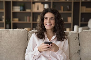 Akıllı telefonuyla kanepede oturmuş gülümseyen çekici bir kadın mutlu görünüyor, internette boş zaman geçiriyor, internette eğleniyor. Genç nesil modern teknolojiyi eğlence ve iletişim için kullanıyor.