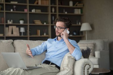 Cep telefonu tutan, konuşma yapan, dizüstü bilgisayarla koltukta oturan, evden çalışan, kanepede dinlenen, projeyi tartışan, satış sonuçlarıyla iş arkadaşlarıyla veya müşterilerle konuşan, bilgi sağlayan, bilgisayar ekranına bakan bir adam.
