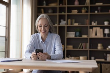 Mutlu yaşlı bayan evde internet uygulamalı akıllı telefon kullanıyor, video görüşmeleri yapıyor, sohbet ediyor, daktilo ediyor, internetten alışveriş yapıyor. Üst düzey cep telefonu kullanıcısı ekrana bakıyor, gülümsüyor, gülüyor