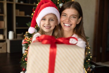 Mutlu, genç bir anne ve küçük bir kız kucağında, Noel hediyesi paketi gösteriyor, dişlek gülümsemeyle kameraya bakıyor, parlayan Xmas ağacına poz veriyor. Şenlikli anne ve kız portresi, yakın çekim.