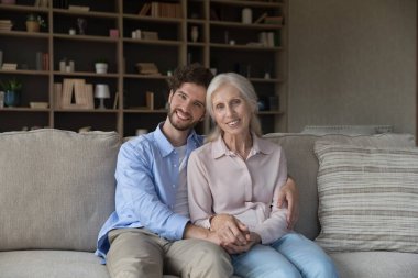 Büyümüş evlat, yaşlı annesine sarılıyor oturma odasında rahat bir kanepede oturuyor poz veriyor, gülümsüyor ve kameraya bakıyor. Mutlu çok nesilli aile portresi, ahenkli ilişkiler, anlayış