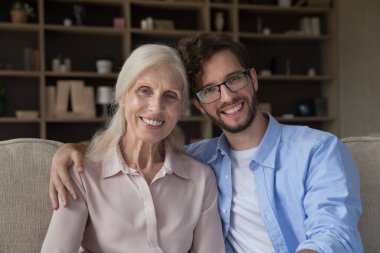 Çekici genç adam oturma odasında kanepede oturur yaşlı anneye sarılır kameraya bakar, poz verir, birlikte boş zamanların tadını çıkarır şefkat, sevgi ve özen gösterir. Yetişkin oğul ve yaşlı annenin aile portresi.