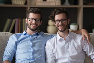 Genç yetişkin ikiz kardeşler, kucaklaşıyorlar, oturma odasında kanepede birlikte oturuyorlar, gülümsüyorlar, kameraya bakıyorlar, arkadaşça iletişimin tadını çıkarıyorlar, birlik oluyorlar, iyi yakın ilişkiler kuruyorlar. Aile bağları, bağ