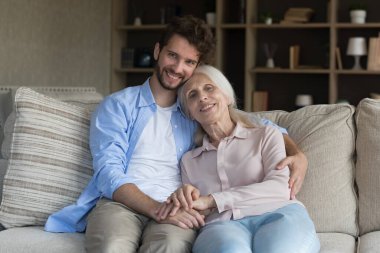 Gülümseyen yetişkin evlat, büyük anne ile rahat bir kanepeye oturur, oturma odası pozunun tadını çıkarır, kameraya bakar, yakışıklı adam büyük anneyi kucaklar sevgi ve özen gösterir. Aile ilişkisi