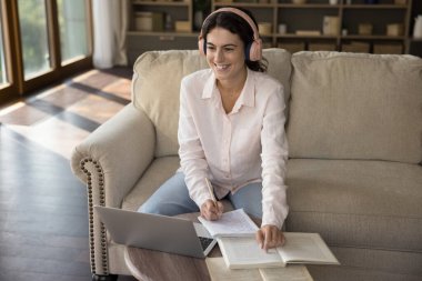 Evde okuyan genç bir bayan öğrenci, kablosuz kulaklık takıyor, defterine notlar yazıyor, ödev yapıyor, dizüstü bilgisayarla üniversite sınavlarına hazırlanıyor. Kişisel eğitim, video çağrı etkinliği, e-öğrenme kavramı