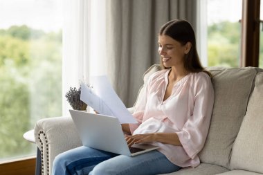 Kağıtları düzenleyen, faturaları yöneten, e-banka uygulaması aracılığıyla online ödemeleri yapan çekici bir kadın kanepeye oturmuş tatmin olmuş görünüyor. Çalışma süreci için internet kullanın, kompozisyon hazırlayın. Teknoloji kavramı