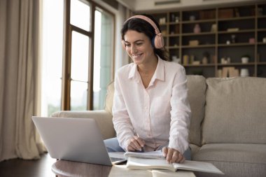 Öğrenci, modern teknoloji kavramını kullanarak evde çalışıyor. Genç kadın rahat oturma odasındaki kanepede oturur dizüstü bilgisayara bakar etkin online sınıf etkinliğinin tadını çıkarır, sesli eğitim kursunun gülümsemesini dinler.