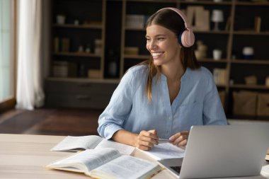 E-öğrenme, eğitim, ses kursu, teknoloji konsepti. Genç, güzel, gülümseyen bir kadın modern kulaklıklar takıyor, çalışıyor, sınavlara hazırlanıyor ders kitapları ve dizüstü bilgisayarlarla masaya oturuyor, yeni bilgiler ediniyor, internet kullanıyor.