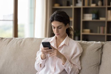 Genç, ciddi bir kadın, elinde akıllı telefonuyla kanepede oturuyor. İnternette sohbet ediyor, mesajlarını paylaşıyor, arama bilgilerini paylaşıyor, web sayfasına bakıyor, internette sörf yapıyor. Yaşam tarzı, modern teknoloji aşırı kullanım kavramı