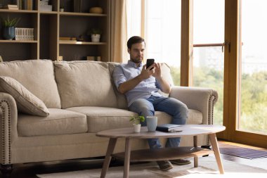 Odaklanmış milenyum adamı oturma odasındaki kanepede rahatlar telefon çalar uygulamasında müzik dinler ve kablosuz kulaklık takar. Kulaklıklı genç adam hafta sonları cep telefonundan video izliyor.