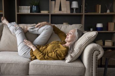 Mutlu, düşünceli, gevşek, gri saçlı kadın rahat koltukta uzanıyor, bacaklarına yaslanıyor, pencereye bakıyor, düşünüyor, hayal ediyor, gülümsüyor. Ev eğlencesi, iyileşme konsepti