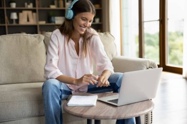 Evde okuyan güzel bir kız öğrenci, dizüstü bilgisayar kullanıyor, kulaklık takıyor, müzik dersi dinliyor, gülümsüyor, rahatına bakıyor, etkin bir şekilde e-öğreniyor. Okul harcı, öğretmenle video görüşmesi, çevrimiçi yeni bilgi kavramı.