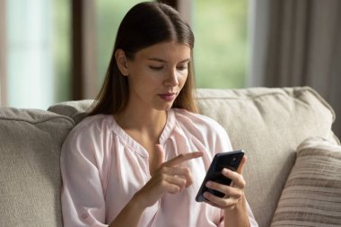 Genç ve güzel bir kadın oturma odasında modern akıllı telefon kullanarak kanepede oturur, evde telefon eder ya da sipariş verir. E-ticaret hizmetleri müşterisi, mobil uygulama kullanımı. Teknoloji, bağlantı, yaşam tarzı konsepti