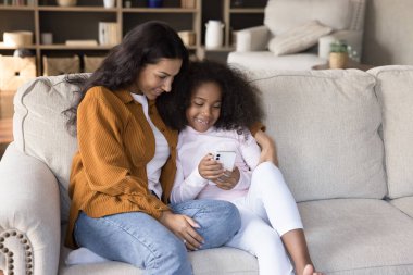 Küçük tatlı Afrikalı kız, genç annesi kanepede dinleniyor, akıllı telefon kullanıyor, yeni video bloğu izliyor, güzel online oyunların tadını çıkarıyor, evde birlikte vakit geçiriyorlar. Teknoloji, cihaz aşırı kullanımı, uygulama kullanımı kavramı