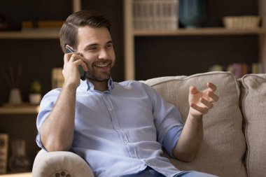 Rahatlamış milenyum erkek işadamı öğrenci, kulağına akıllı telefon tutarak güzel bir telefon görüşmesi yapıyor. Mutlu genç erkek sohbeti evdeki kanepede otururken telefonda konuşuyor.