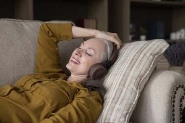 Cheerful peaceful elderly woman wearing modern wireless headphones listening to music, enjoying songs, entertainment, lazy break, leisure time, chill, lying on couch at home, smiling, laughing clipart