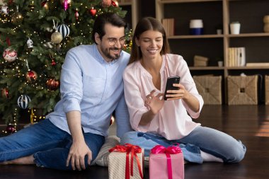 Mutlu milenyum çiftleri süslü Noel ağacında dinleniyor, hediye kutuları, ekommerce uygulamasını akıllı telefonlarda kullanıyor, internetten alışveriş yapıyor, internette sohbet ediyor, Mutlu Noeller mesajı yolluyor.