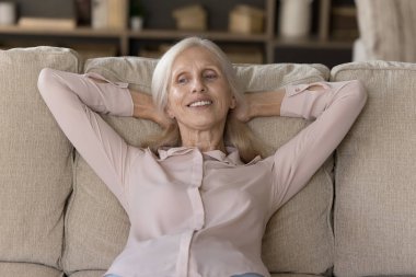 Huzurlu, yaşlı, gülümseyen kadın ellerini başının arkasına koy rahat kanepeye uzan evde tek başına dinlen, temiz havanın tadını çıkar rahat oturma odasında, rahatla, kaygısız, emekli bir hayat yaşa.