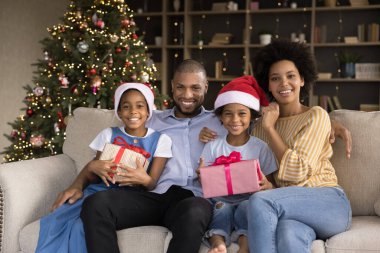 Mutlu siyah aile çiftleri ve yeni yılı evde kutlayan kardeş çocuklar. Noel ağacındaki kanepede dinlenen Noel Baba şapkalı küçük aileler ve çocuklar ellerinde Noel hediyesi kutuları, gülümsüyorlar. Kutlama portresi