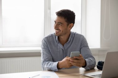 Yakışıklı İspanyol işadamı iş yerinde dikkati dağılmış vaziyette oturuyor elinde akıllı telefon tebessümü, yana bakıyor, iş için mobil uygulamalar, rahat iş akışı modern kablosuz teknoloji kavramı kullanıyor