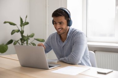 İspanyol genç işadamı, kulaklıklı ofis çalışanı iş yerinde dizüstü bilgisayarla oturuyor, internette çalışıyor, video çağrı uygulamasında konuşuyor. İş dünyası modern kablosuz teknoloji ve sanal toplantı kavramını kullanır