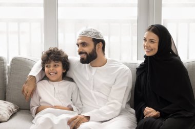 Mutlu Arap ebeveynler, sevimli oğlanı kucaklıyor, evdeki kanepede aile fotoğrafı için poz veriyorlar. Geleneksel giysiler içinde, gülümseyen, gülen, gülümseyen Müslüman bir çift ve küçük bir çocuk.