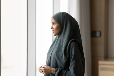 Gri başörtüsü ve abaya elbisesi giymiş düşünceli genç Arap iş kadını büyük ofis penceresinden dışarı bakıyor, başarılı bir kariyer ve iş planı düşünüyor. Yan görünüm portresi