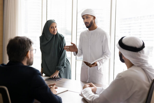 Couple of young Arabian managers in Muslim clothes presenting project to European business partners, speaking to colleagues sitting at meeting table, discussing company selling, investment