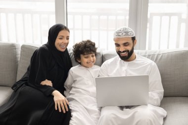 Neşeli Arap anne, baba ve yakışıklı oğlan müslüman kıyafetleri içinde multimedya uygulaması, dizüstü bilgisayarda internet servisi, birlikte kanepede oturup, film, dizi, gülücük, gülücük