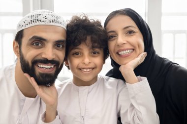 Neşeli Müslüman anne-baba ve oğul Arap kıyafetleri içinde evde yakın çekim portresi için poz veriyor, kameraya bakıyor, gülümsüyor, gülüyor. Mutlu şirin çocuk anne ve babasının yüzüne dokunuyor.