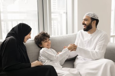 Geleneksel giysiler içinde neşeli Arap ebeveynler evdeki kanepede oğlunu gıdıklıyor, sevimli çocuğa sarılıyor, etkinliğin tadını çıkarıyor, aile eğlencesi, gülüyor, kıkırdayan çocukla eğleniyor.