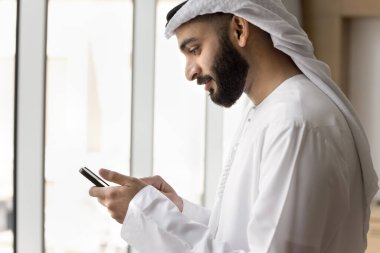Beyaz Müslüman erkek giysileri içinde, iş için kablosuz internet iletişimini kullanan, akıllı telefon kullanan, sosyal medyada gezinen, internette sohbet eden genç, pozitif Arap iş adamı. Yan görünüm