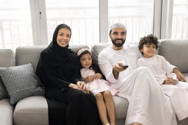 Mutlu Müslüman ebeveynler ve iki kardeş çocuk film seyrediyor, aile dizisi, uzaktan kumanda kullanıyor, kanal değiştiriyor, düğmeye basıyor, kanepede oturuyor, gülümsüyor, gülüyor.