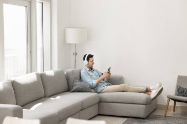 Tembel, rahat, kulaklıklı, evde rahat bir koltukta dinlenen, müzik dinleyen, online müzik listesindeki melodilerden hoşlanan, akıllı telefondaki multimedya servisini kullanan genç bir Latin. Tam vücut çekimi.