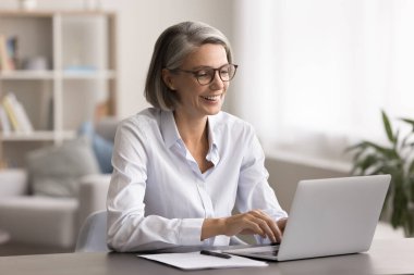 Gülümseyen orta yaşlı kadın masamda oturup dizüstü bilgisayara, kablosuz internete bakıyor, çevrimiçi projeye hazırlanıyor, iş arkadaşlarıyla sohbet ediyor, uzaktan işten zevk alıyor, modern teknolojiyi e-ticaret için kullanıyor.